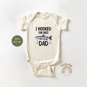 May include: A cream-colored baby onesie with a fishing-themed design. The onesie says "I hooked the best dad" with a cartoon fish and fishing hooks.