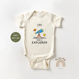 May include: Cream-colored baby onesie with a cartoon graphic of a blue telescope, planet, and star. The text reads "TINY SPACE EXPLORER". The onesie is made of soft material.