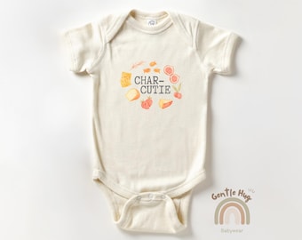 May include: Cream-colored baby onesie with the text "CHAR-CUTIE" in a playful font, surrounded by watercolor illustrations of cheese, figs, and strawberries. The onesie has short sleeves and a snap closure. The brand logo "Gentle Hug Babywear" is at the bottom.
