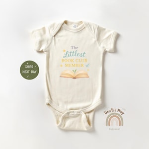 May include: Cream-colored baby onesie with the text "The Littlest Book Club Member" and an illustration of an open book. The design includes stars and a small plant growing from the book. The onesie is a short-sleeved style.