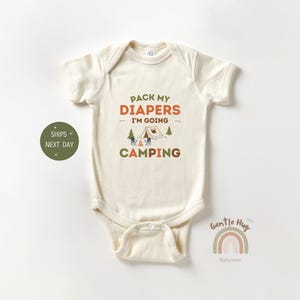 May include: Cream-colored baby onesie with the text "PACK MY DIAPERS I'M GOING CAMPING" in orange and green. The design includes a tent, trees, and a campfire illustration. A rainbow logo and the words "Gentle Hug Babywear" are also visible.