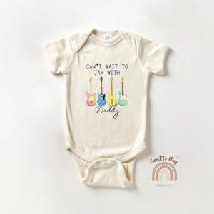 May include: Cream-colored baby bodysuit with the text "CAN'T WAIT TO JAM WITH Daddy" and four colorful electric guitars. The guitars are pink, blue, yellow, and green. A rainbow and the words "Gentle Hug Babywear" are at the bottom.
