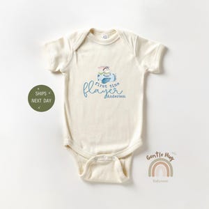 May include: A cream-colored baby onesie with a blue airplane graphic and the text "first time flyer Anderson".