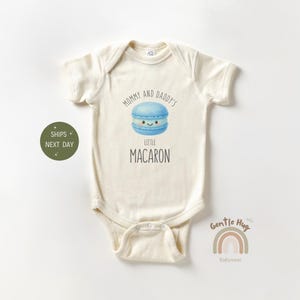 May include: Cream-colored baby bodysuit with short sleeves. The front features a blue watercolor illustration of a macaron with a smiling face, and the text "Mommy and Daddy's Little Macaron". A green circle with text is on the left.