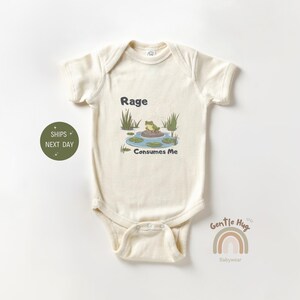 May include: A cream-colored baby onesie with short sleeves features a green frog sitting on a lily pad in a pond. The text "Rage Consumes Me" is printed below the frog. The onesie is from the brand "Gentle Hug Babywear."