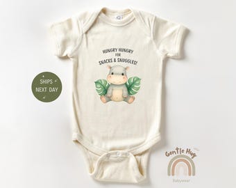 May include: Cream-colored baby onesie with a cute hippo illustration and the text "HUNGRY HUNGRY FOR SNACKS & SNUGGLES!". The hippo is gray with green leaves on either side. Gentle Hug Babywear logo is at the bottom.