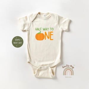 May include: Cream-colored baby bodysuit with the text "HALF WAY TO ONE" in green and orange. A cartoon pumpkin graphic is between the words "TO" and "ONE". The bodysuit has short sleeves and a snap closure.