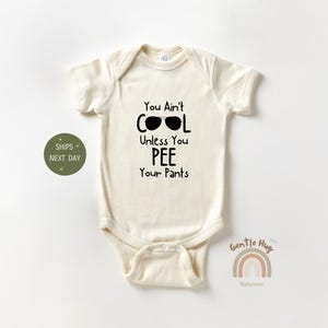 May include: A white baby onesie with black text that reads "You Ain't Cool Unless You Pee Your Pants" with a pair of sunglasses above the word "Cool".