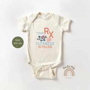 May include: A cream-colored baby onesie with a graphic of colorful pills and the text "Your Rx of Cuteness is Filled".
