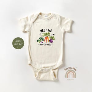 May include: A cream-colored baby onesie with the text "Meet me at the farmer's market" in black. The onesie features a colorful illustration of vegetables, including carrots, broccoli, and a beet.