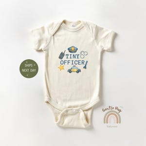 May include: Cream-colored baby onesie with a "Tiny Officer" design. The design includes a police hat, handcuffs, a walkie-talkie, a star, a police car, and a flashlight. The onesie is made of soft material.
