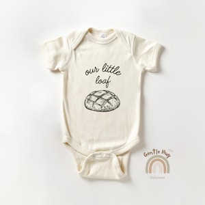 May include: Cream-colored baby onesie with short sleeves. The front features the text "our little loaf" above a detailed illustration of a loaf of bread. The bottom right corner has a rainbow graphic and the text "Gentle Hug Babywear."