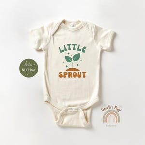 May include: A cream colored baby onesie with the text "Little Sprout" in green and orange letters. A green sprout with two leaves is above the text. The onesie is from the brand "Gentle Hug Babywear".