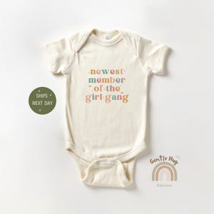 May include: A cream-colored baby onesie with the text "newest member of the girl gang" in colorful letters. The onesie is a short-sleeved bodysuit with a snap closure.
