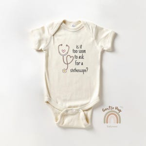 May include: Cream-colored baby bodysuit with a stethoscope graphic and the text "is it too soon to ask for a stethoscope?" The design includes a heart and flower. The brand logo "Gentle Hug Babywear" is at the bottom.
