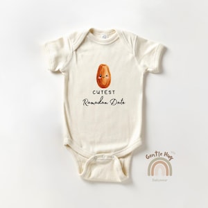 May include: Cream-colored baby onesie with a cute cartoon date fruit illustration and the text "Cutest Ramadan Date". The onesie has short sleeves and a snap closure at the bottom. The brand logo "Gentle Hug Babywear" is at the bottom right.