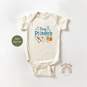 May include: Cream-colored baby onesie with a blue and red "Tiny Plumber" graphic. The design includes a wrench, faucet, toolbox, and other plumbing tools. The onesie is made of soft material.