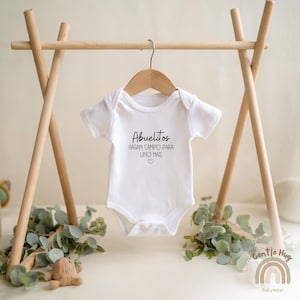 K&ouml;nnte beinhalten: Wei&szlig;er Baby-Strampler mit schwarzem Text, der "Abuelitos hagan campo para uno mas" mit einem kleinen Herzsymbol lautet.