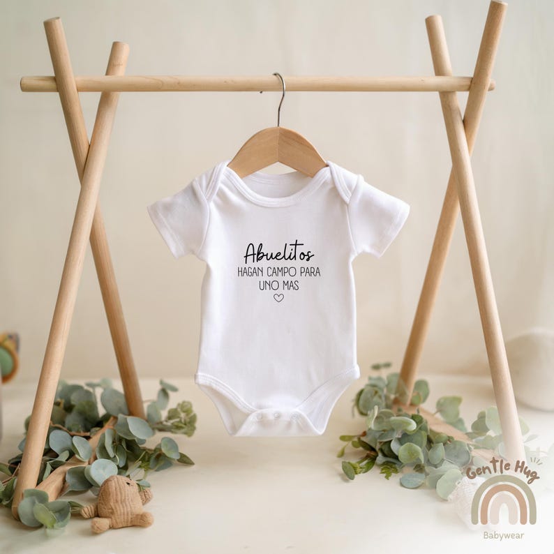 K&ouml;nnte beinhalten: Wei&szlig;er Baby-Strampler mit schwarzem Text, der "Abuelitos hagan campo para uno mas" mit einem kleinen Herzsymbol lautet.