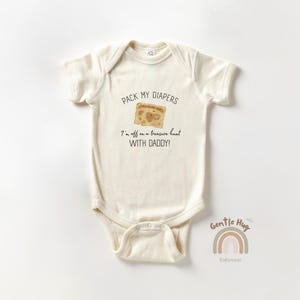 May include: Cream-colored baby onesie with short sleeves. The front features the text "PACK MY DIAPERS" above a treasure map graphic. Below, it reads "I'm off on a treasure hunt WITH DADDY!" A rainbow graphic and the words "Gentle Hug Babywear" are at the bottom.