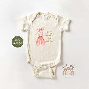 May include: Cream-colored baby onesie with short sleeves. The front features a watercolor illustration of pink ballet shoes tied with a pink ribbon. Text reads "Tiny Dancer Big Moves" in brown. A green circle with text is on the left.