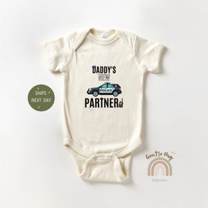 May include: A cream-colored baby onesie with a black and white graphic of a police car and the text "Daddy's New Partner".