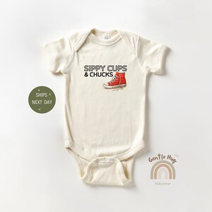 May include: Cream-colored baby onesie with the text "SIPPY CUPS & CHUCKS" and a red sneaker graphic. The onesie is a soft, comfortable garment for infants. The design is playful and suitable for a baby.