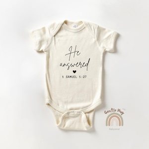 May include: Cream-colored baby onesie with the text "He answered" in a handwritten font, a small black heart, and the scripture reference 1 Samuel 1:27. The onesie has short sleeves and a snap closure.