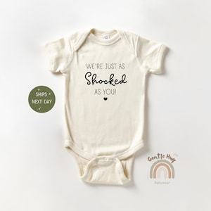 May include: Cream-colored baby bodysuit with short sleeves. The text "WE'RE JUST AS SHOCKED AS YOU!" is printed in black, with a small heart below. A green circle with text is on the left. A rainbow logo is on the right.
