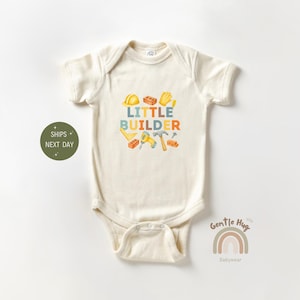 May include: Cream-colored baby onesie with the text "LITTLE BUILDER" in colorful letters. The design includes illustrations of a yellow hard hat, bricks, gloves, a hammer, and other construction tools. A rainbow and the words "Gentle Hug Babywear" are also visible.