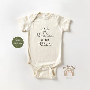 May include: Cream-colored baby onesie with the text "Cutest Pumpkin in the Patch" in black script. A small pumpkin graphic is above the word "Pumpkin". The onesie has short sleeves and a snap closure.
