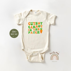May include: A cream-colored baby onesie with the text "Cutest Carrot in the Patch" in green and orange letters. A small orange carrot with a smiling face is next to the text.