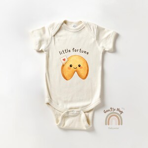 May include: Cream-colored baby bodysuit with a cartoon fortune cookie graphic. The fortune cookie has a smiling face and a small heart. The text "little fortune" is above the cookie. The brand name "Gentle Hug Babywear" is below the graphic.