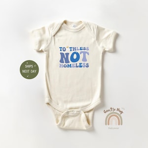 May include: A white baby onesie with a blue and purple graphic that says "Toothless Not Homeless". The onesie is a short-sleeved bodysuit with a snap closure at the bottom.