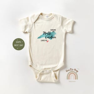 May include: Cream-colored baby bodysuit with the text "Someone in North Carolina Loves Me" and a teal map of North Carolina. Two small red hearts are below the text. The bodysuit has short sleeves.