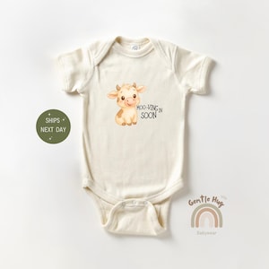 May include: Cream-colored baby onesie featuring a cartoon cow illustration with the text "MOO-VING IN SOON." The onesie has short sleeves and a snap closure. The design includes a smiling cow with brown spots and small horns.