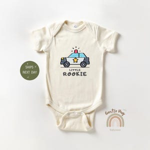 May include: A cream-colored baby onesie with a cartoon police car and the text "Little Rookie".