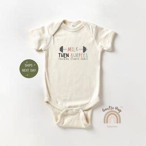 May include: Cream-colored baby onesie with the text "MILK THEN BURPEES TRAINING STARTS EARLY" in a playful font. The design includes a barbell graphic. A small rainbow graphic and the words "Gentle Hug Babywear" are also visible.