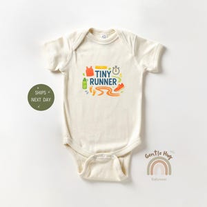 May include: Cream-colored baby onesie with a colorful graphic that reads "Tiny Runner." The design includes a stopwatch, running shoes, a water bottle, and a running track. The onesie is made of soft material.