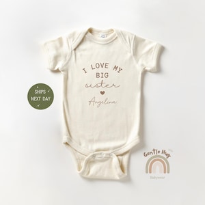 May include: Cream-colored baby bodysuit with short sleeves. The text "I LOVE MY BIG sister" is printed in brown, with a heart symbol and the name "Angelina" below. The bodysuit has a snap closure.