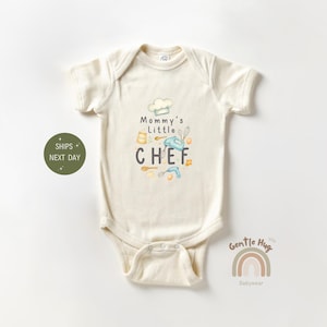 May include: Cream-colored baby onesie with the text "Mommy's Little Chef" and illustrations of cooking tools. The design includes a chef's hat, mixer, and measuring spoons. The onesie is made of soft material.