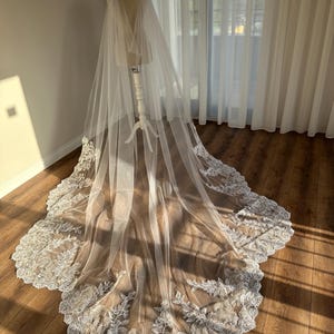 May include: A white tulle wedding veil with a scalloped lace trim. The veil is draped over a mannequin and has a cape-like design.