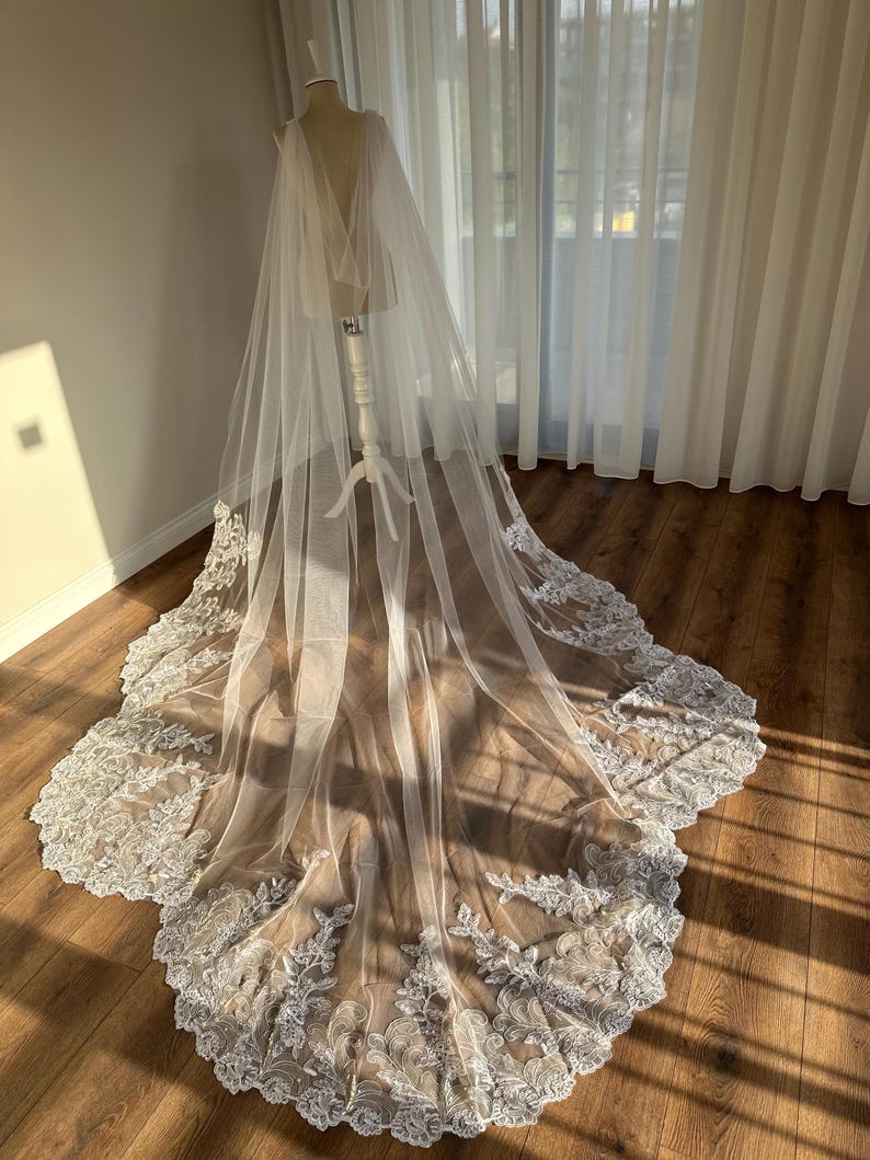 May include: A white tulle wedding veil with a scalloped lace trim. The veil is draped over a mannequin and has a cape-like design.