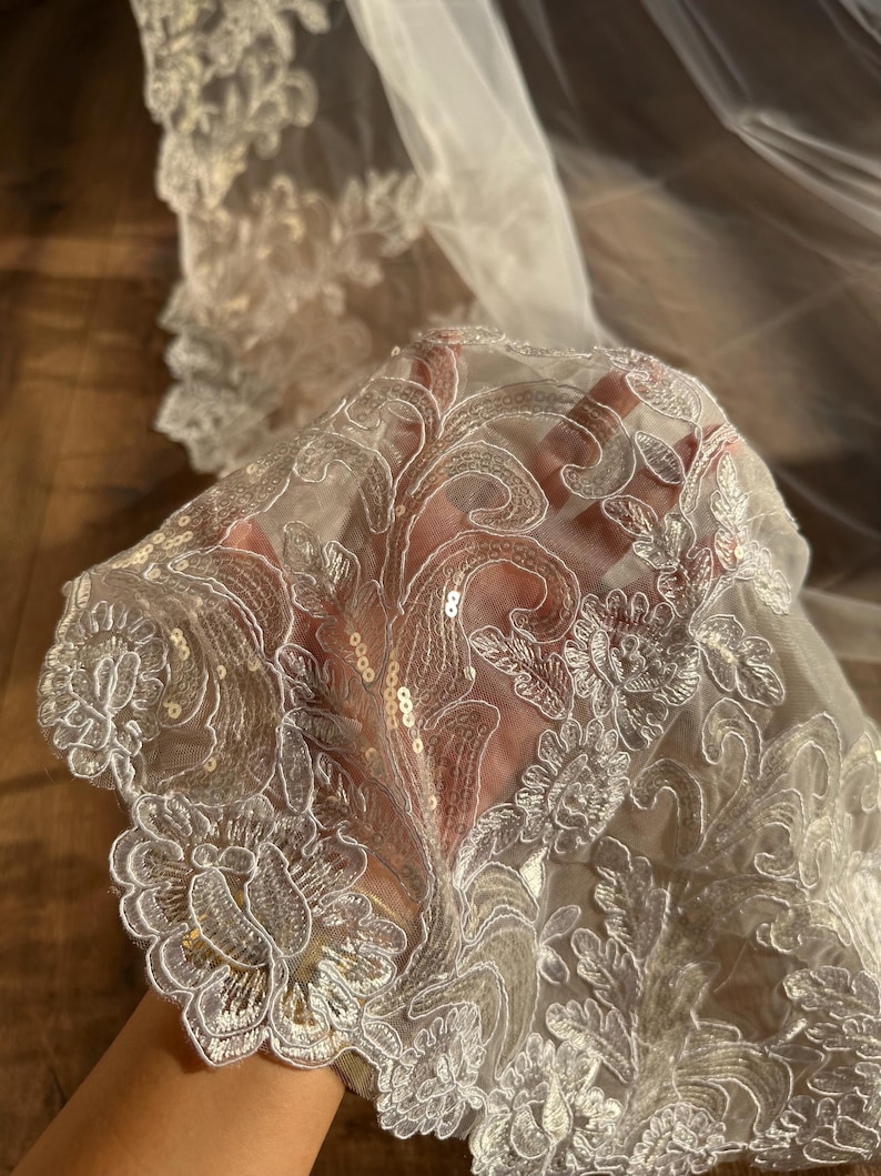 May include: White bridal veil with a delicate floral lace trim and sequins. The lace is intricate and features a floral pattern.