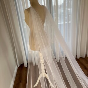 May include: A white tulle bridal cape draped over a white dress form. The cape is sheer and has a flowing, elegant design.