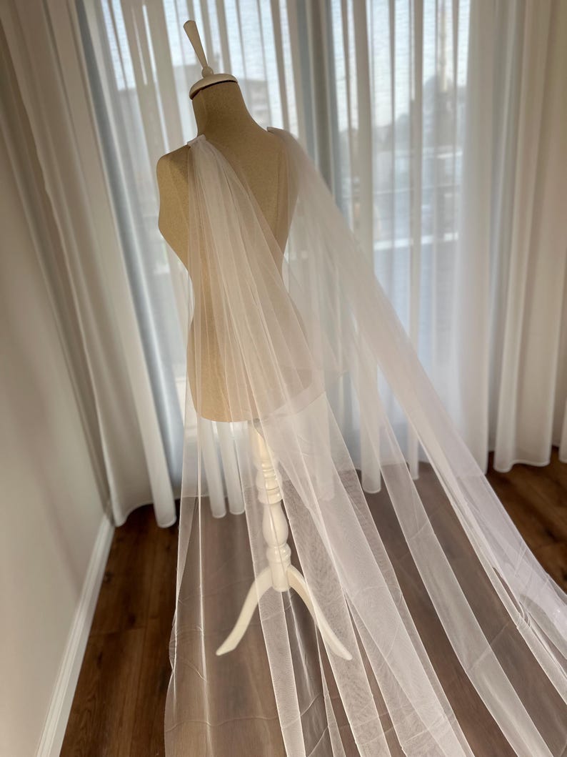 May include: A white tulle bridal cape draped over a white dress form. The cape is sheer and has a flowing, elegant design.