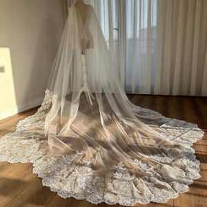 May include: A white tulle wedding veil with a delicate lace trim. The veil is draped over a mannequin and has a long train.