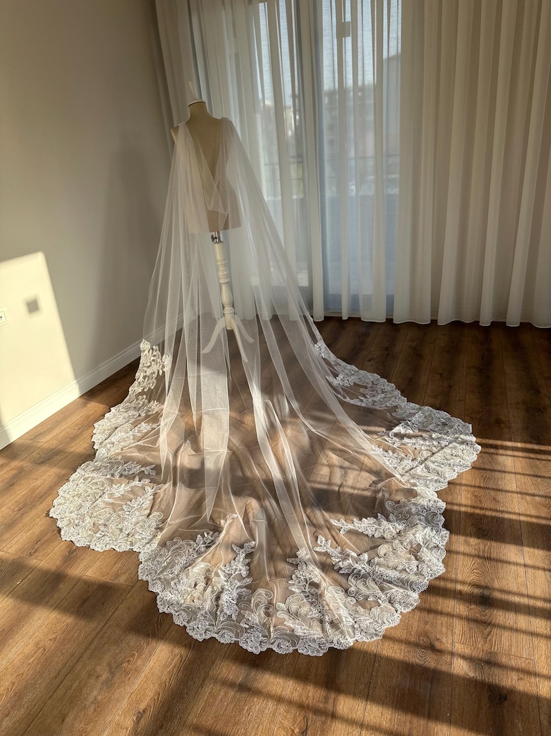 May include: A white tulle wedding veil with a delicate lace trim. The veil is draped over a dress form and has a long train.
