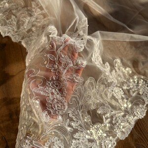 May include: White tulle fabric with a delicate floral lace trim. The lace features a floral pattern with sequins.