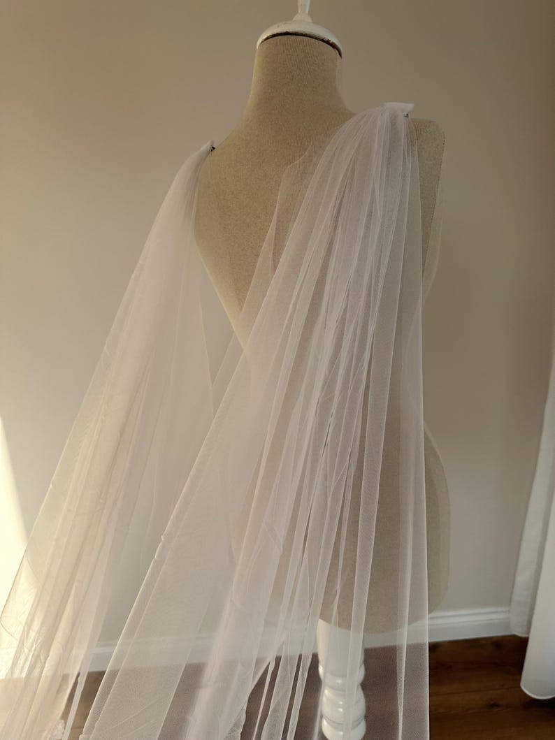 May include: A white tulle wedding veil draped over a dress form. The veil is sheer and delicate, with a soft, flowing texture.